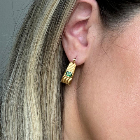 A close-up of a model's ear wearing a gold geometric drop earring with a green rectangular stone.