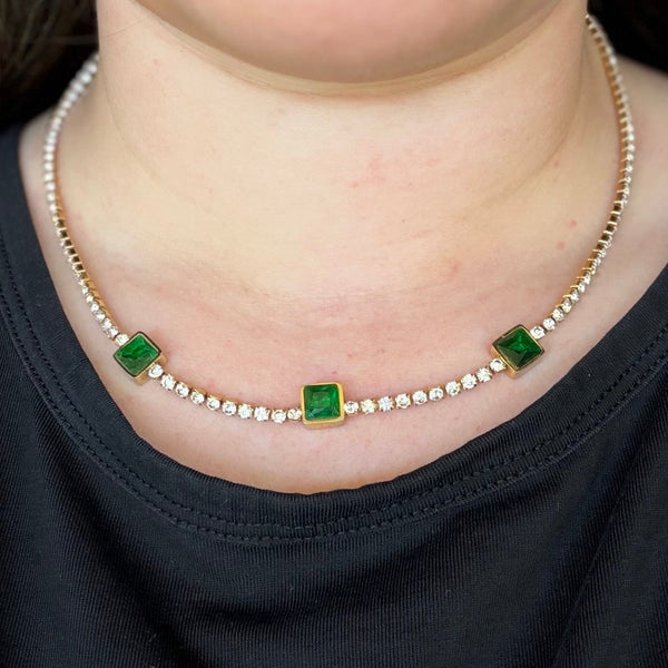 A close-up of a woman wearing a delicate gold chain necklace with three small square green gemstones and a line of tiny clear stones, paired with a black shirt.