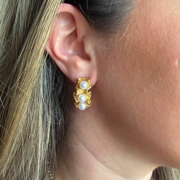 A close-up side profile of a woman wearing a gold huggie hoop earring with three white faux pearls, along with a gold beaded necklace.