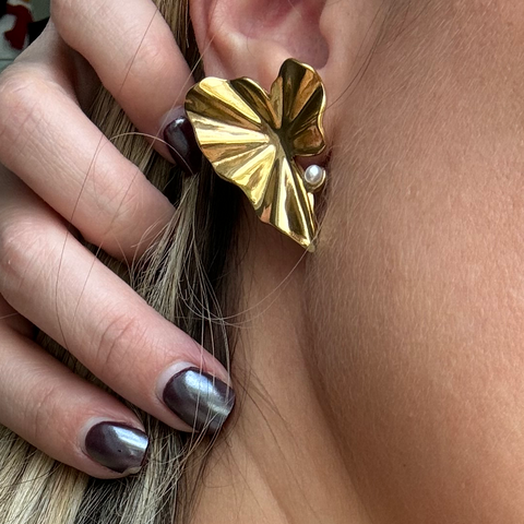 A close-up lifestyle shot of a person wearing a textured gold leaf-shaped earring with a small pearl accent, paired with a pearl necklace. The person has blonde hair with dark roots and metallic gray nail polish.